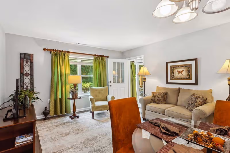A cozy living room with a beige sofa adorned with patterned cushions, a green armchair near a window with green curtains, a wooden side table with a lamp, a glass dining table with orange chairs, and a framed butterfly artwork on the wall.