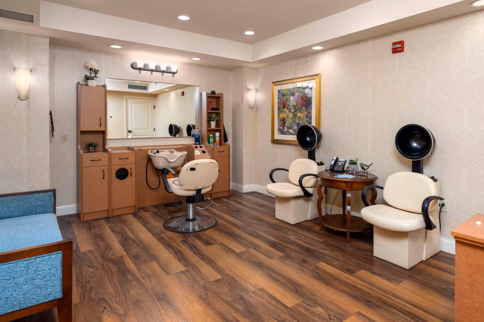 Interior of a salon area in a senior living facility with a hair washing station, two cream-colored salon chairs with black hair dryers, a wooden side table with a phone and decorative items, a large mirror, and a blue cushioned bench. The room has wood flooring and beige patterned wallpaper.