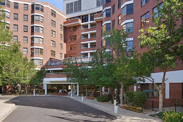 Front exterior of a multi-story red-brick senior living building with a covered entrance, driveway, trees, and landscaping.