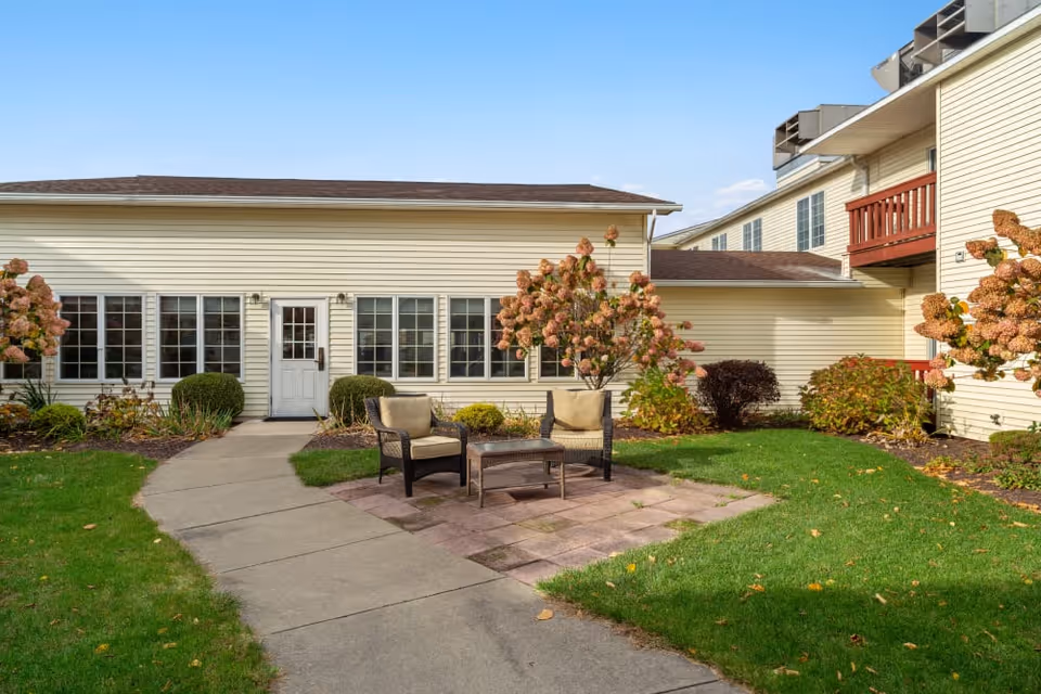 Outdoor patio area at Brookdale Castle Gardens featuring two cushioned chairs and a small table on a paved section surrounded by green grass, flowering bushes, and beige buildings with multiple windows under a clear blue sky.