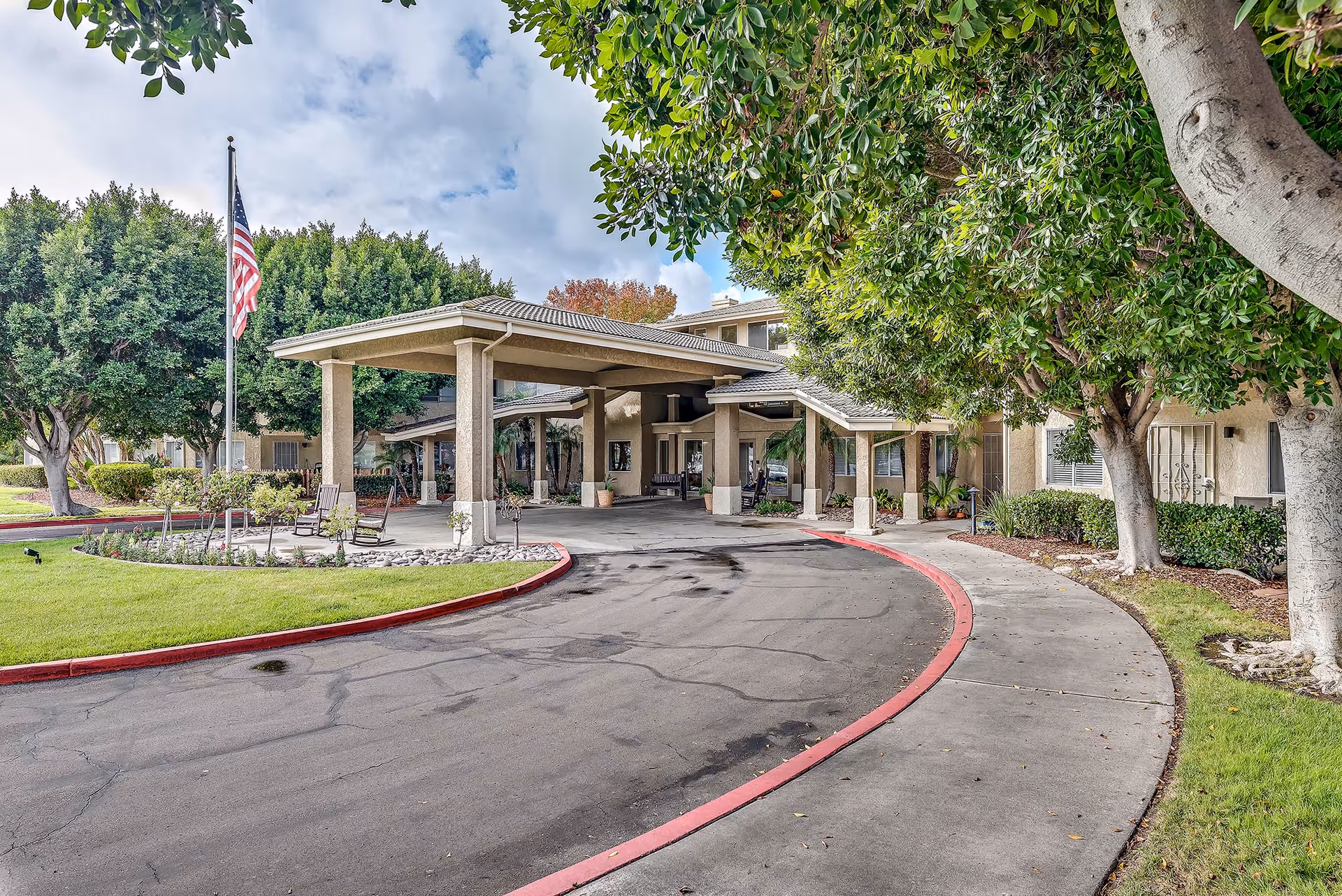 Front exterior view of Arcadia Place Senior Living facility with a covered entrance, an American flag on a flagpole, trees, and a curved driveway bordered by a sidewalk and green lawn.