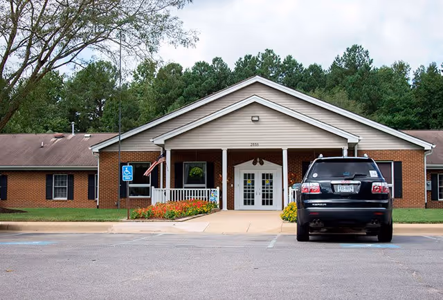Front exterior view of a single-story brick building with a white entrance porch, double glass doors, and a small garden with flowers. A black SUV is parked in a handicapped parking spot in front of the building. Trees and greenery are visible in the background under a cloudy sky.