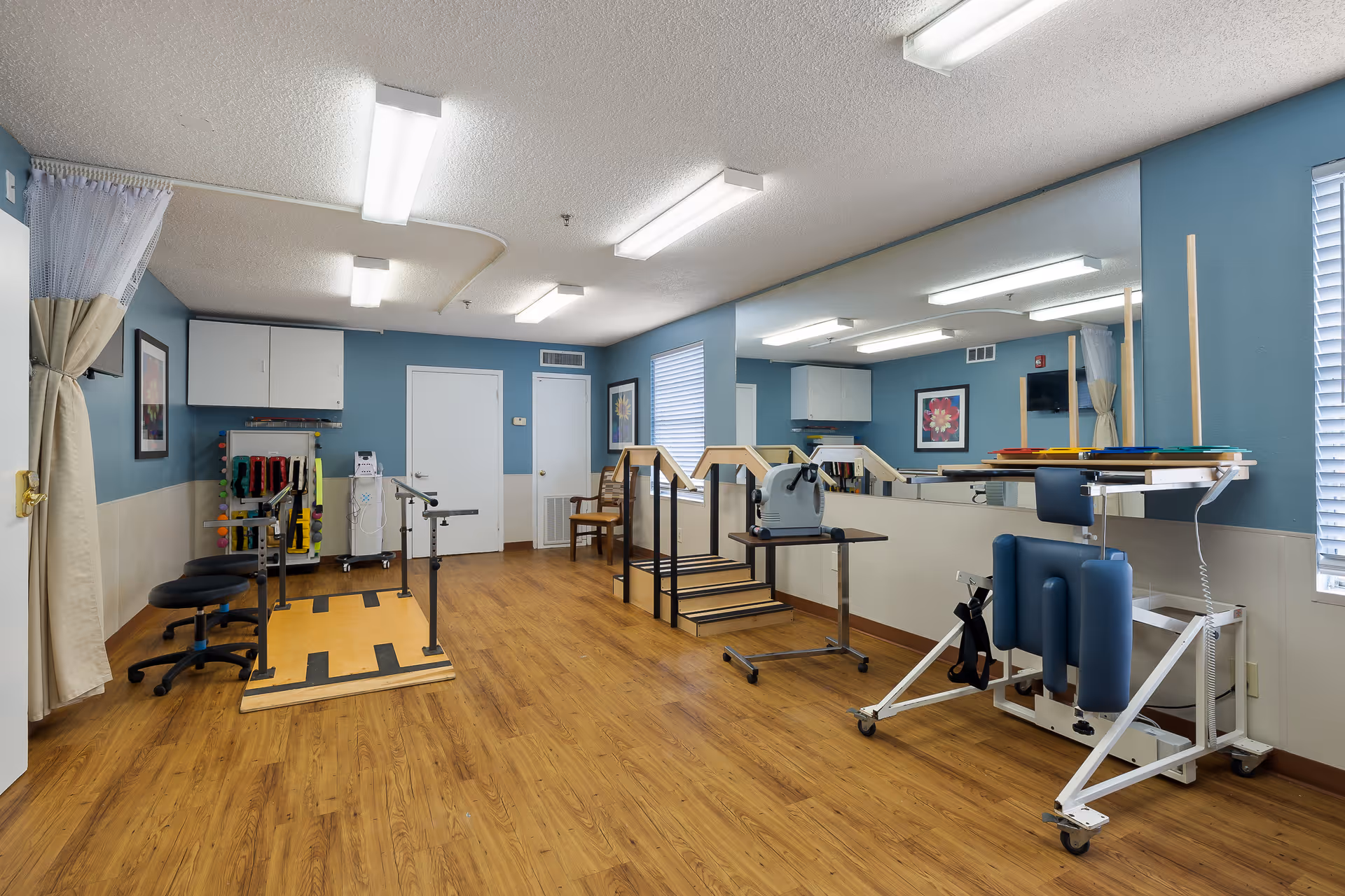 A rehabilitation therapy room with wooden flooring and blue walls. The room contains parallel bars for walking exercises, a small set of stairs with handrails, exercise equipment, chairs, and a large mirror on one wall. The room is well-lit with ceiling lights and has windows with blinds.