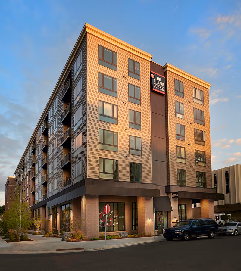 Exterior view of a multi-story senior living building, The Pillars of Prospect Park, on a street corner at sunset.