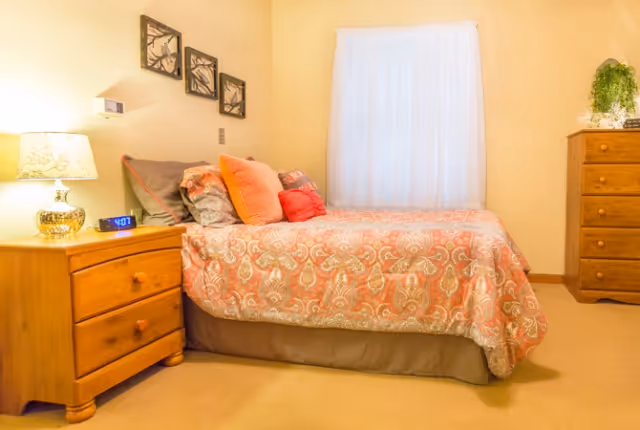 Cozy bedroom featuring a patterned coral bed, wooden nightstand and dresser, lamp, and a curtained window.