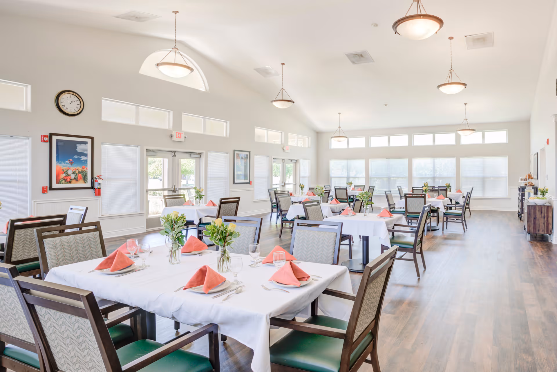 Bright and spacious dining room with multiple tables covered in white tablecloths, each set with pink folded napkins, glassware, and small floral centerpieces. The room has large windows allowing natural light to fill the space, wooden flooring, and several hanging light fixtures on the ceiling.