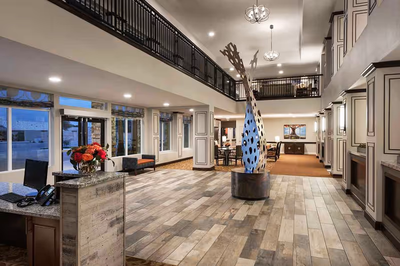 Spacious and well-lit senior living facility lobby with a modern art sculpture in the center, a reception desk with a computer and phone on the left, seating areas with chairs and tables in the background, large windows letting in natural light, and a second-floor balcony overlooking the area.