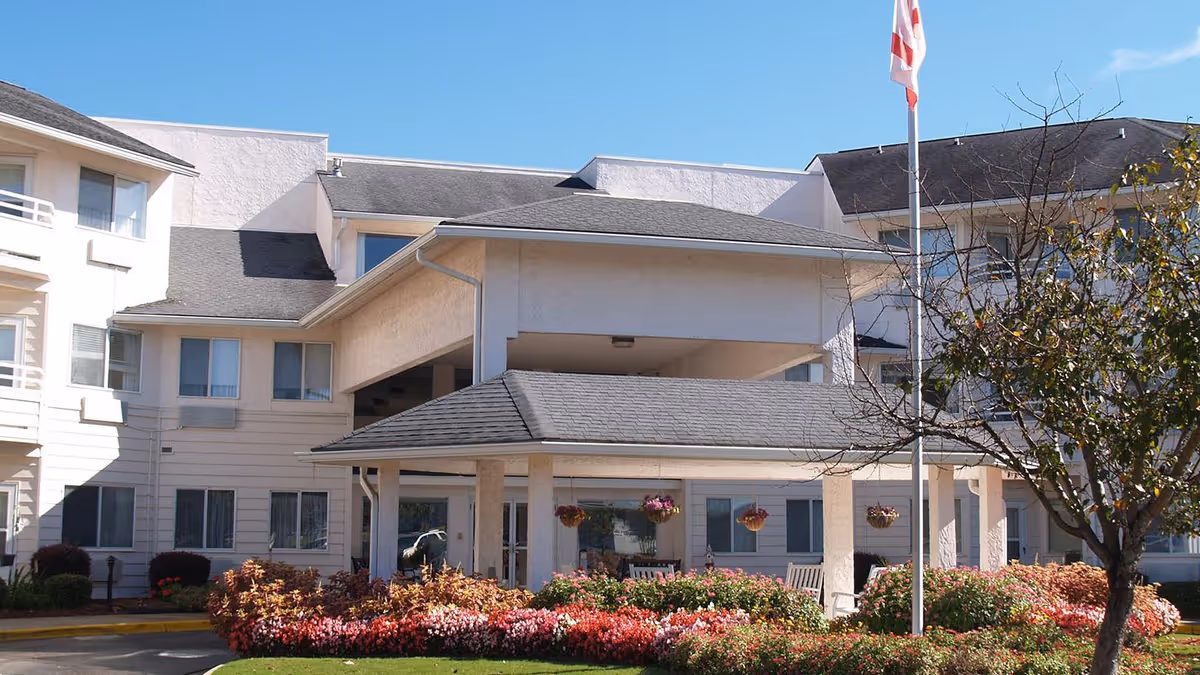 Front entrance of a multi-story senior living building with a covered porte-cochere, flagpole and landscaped flower beds.