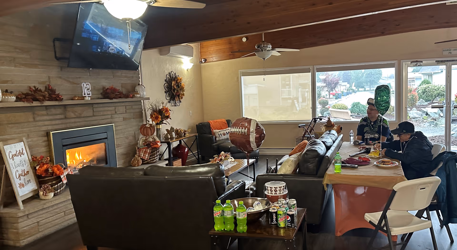 A cozy living room area with a stone fireplace lit with a fire, decorated with autumn-themed items. There are two dark leather sofas, a coffee table with decorations, and a TV mounted above the fireplace. Two people are seated at a table covered with a tablecloth, eating and drinking. Large windows and a glass door provide a view of the outside.