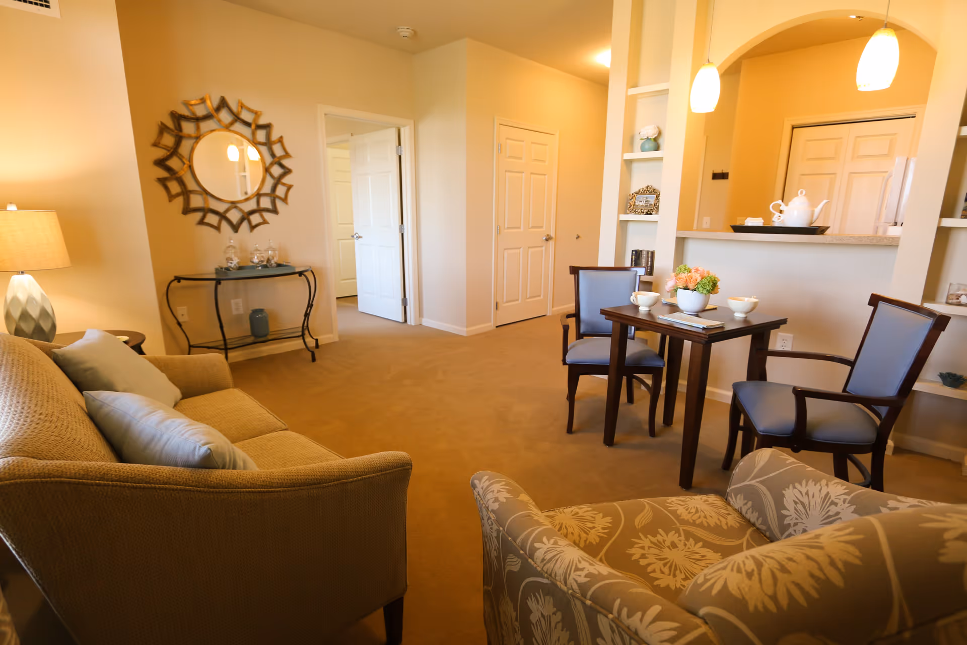 A cozy living room area with a beige sofa, a patterned armchair, and a small wooden table with two chairs. The table has a flower arrangement and two cups on it. There is a decorative mirror on the wall above a small console table with glass jars. The room has beige walls and carpet, with a view into an adjacent kitchen area with pendant lights and a white teapot on the counter.