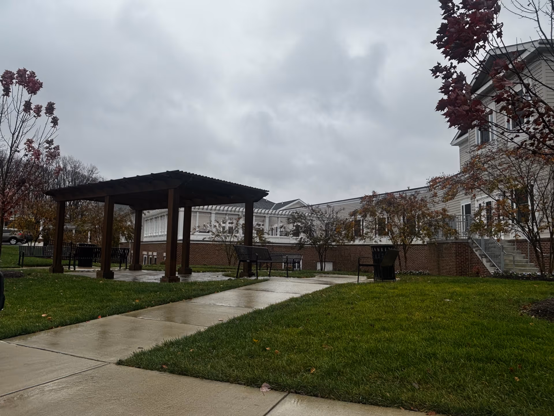 Outdoor area of HarborChase of Olney featuring a wooden pergola with benches underneath, surrounded by green grass and trees with autumn foliage. The building is visible in the background under a cloudy sky.