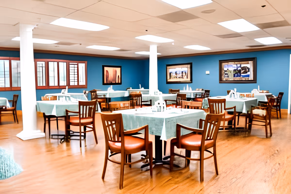 A spacious dining room with multiple tables covered with light blue tablecloths, each surrounded by wooden chairs with armrests. The room has blue walls adorned with framed artwork, large windows, and white ceiling lights. The floor is wooden, and there are white columns in the room.