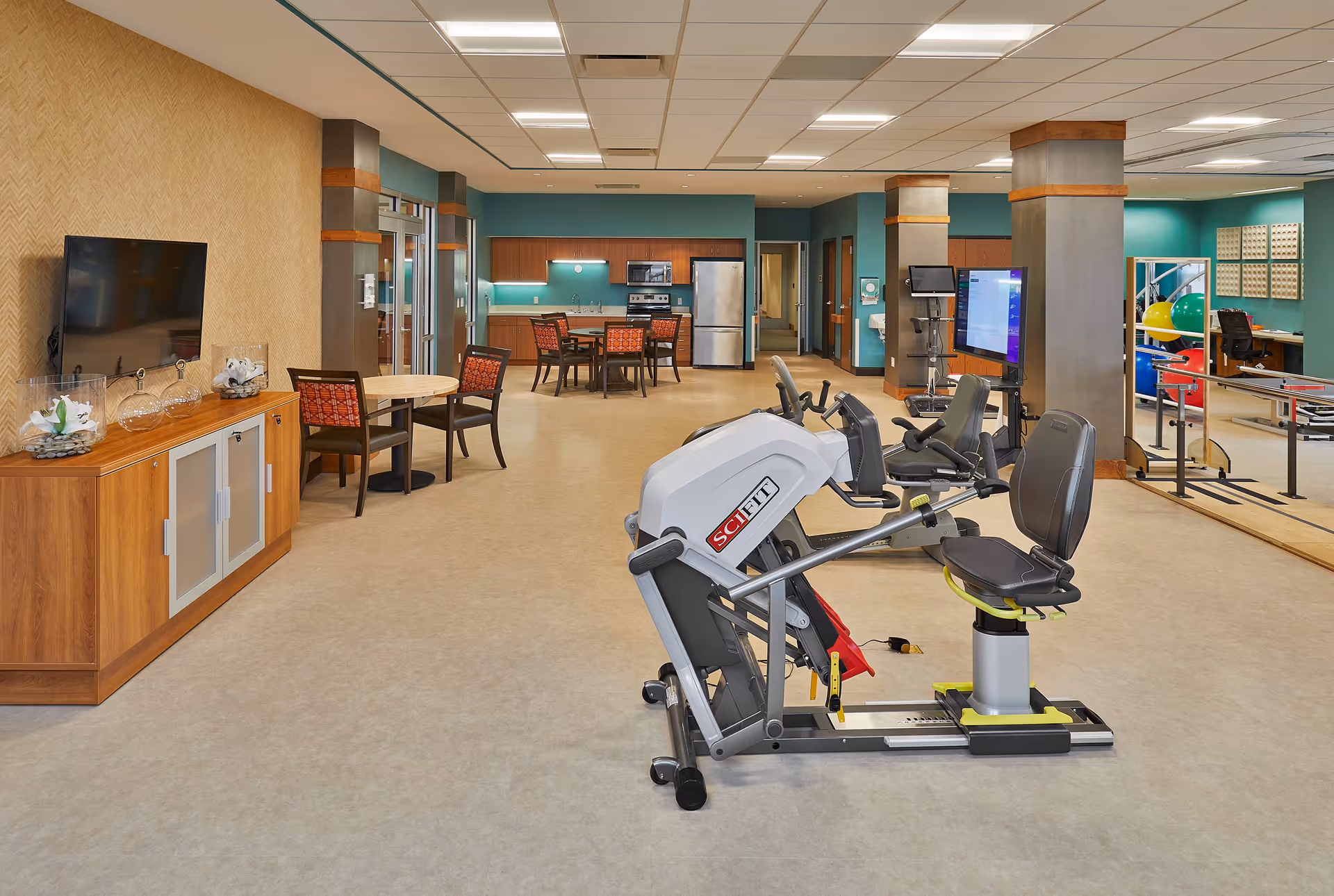 Open interior activity and therapy room with exercise machines in the foreground, seating and a kitchenette in the background.