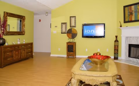 A cozy living room with light green walls, a wooden sideboard with a large mirror above it, a flat-screen TV mounted on the wall, decorative wall art, a fireplace with a mantel, and a coffee table with a decorative bowl on top.