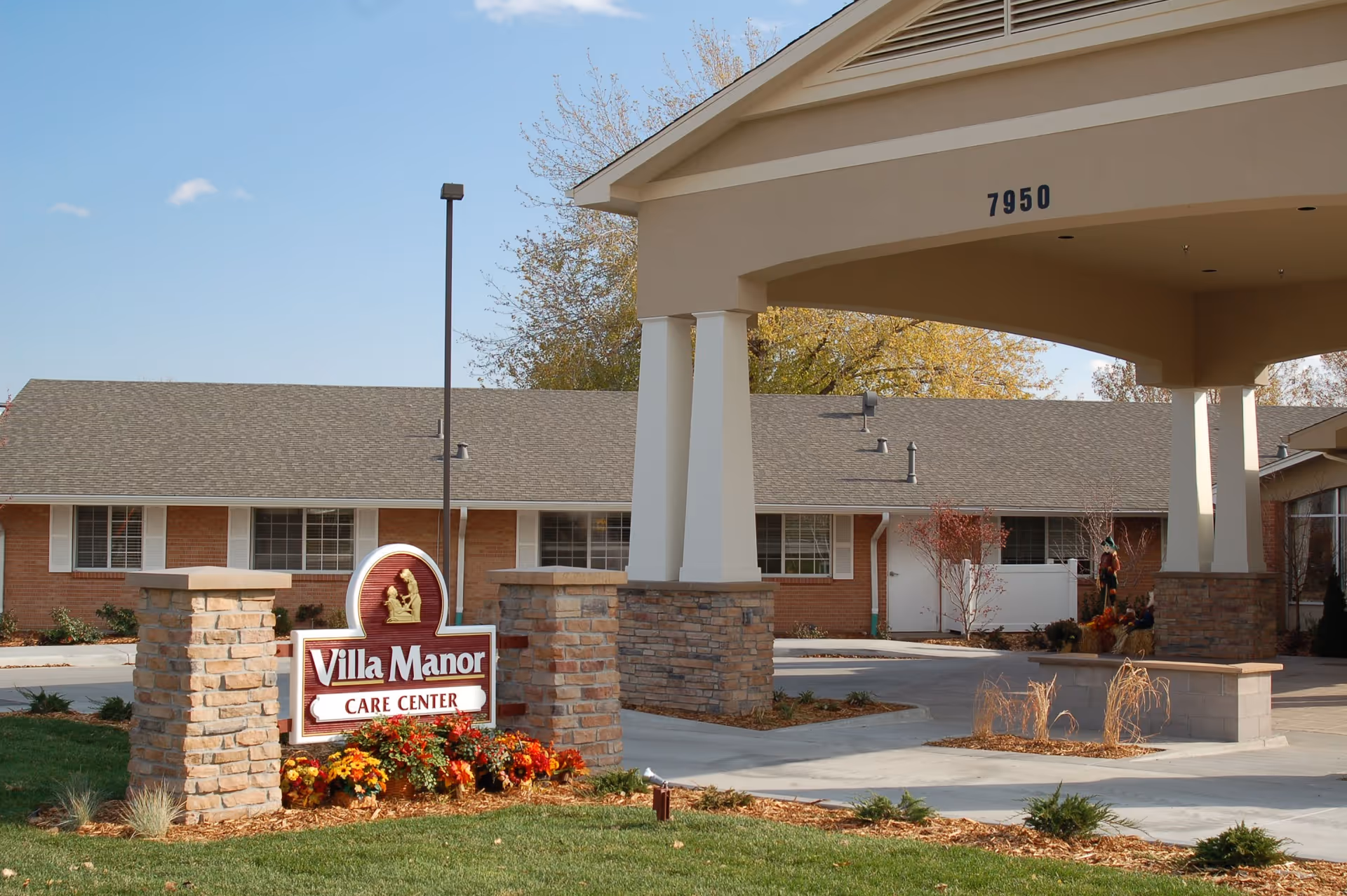 Entrance of Villa Manor Care Center showing a covered drop-off area with pillars and a sign in front surrounded by flowers and landscaping. The building is a single-story brick structure with white window shutters under a clear blue sky.