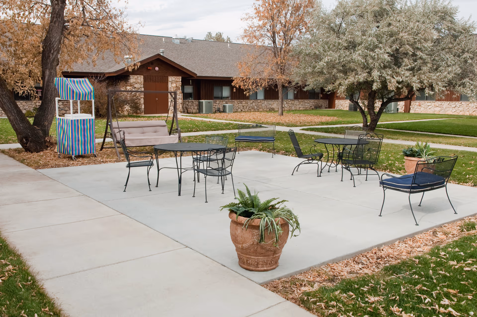 Outdoor courtyard patio with metal tables, chairs, a swing, and potted plants in front of a single-story building.