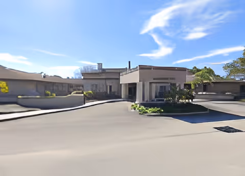 Exterior view of a single-story building under a blue sky with some clouds, featuring a driveway and some greenery near the entrance.