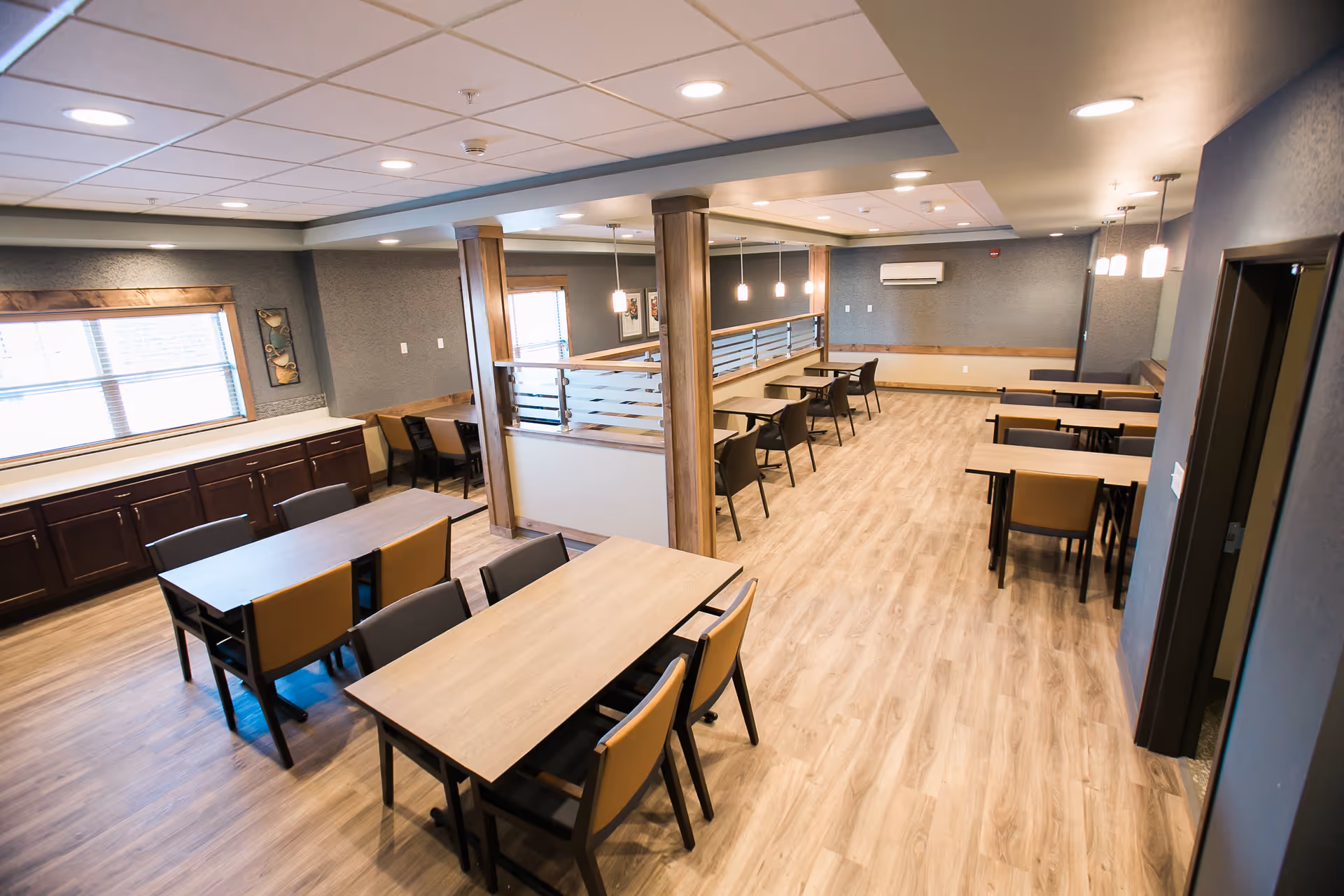 A spacious dining room with multiple wooden tables and chairs arranged neatly on a light wood floor. The room features a modern design with gray walls, wooden trim, and ceiling lights. Large windows with blinds allow natural light to enter. There are wooden pillars and a half wall with frosted glass panels dividing the space.