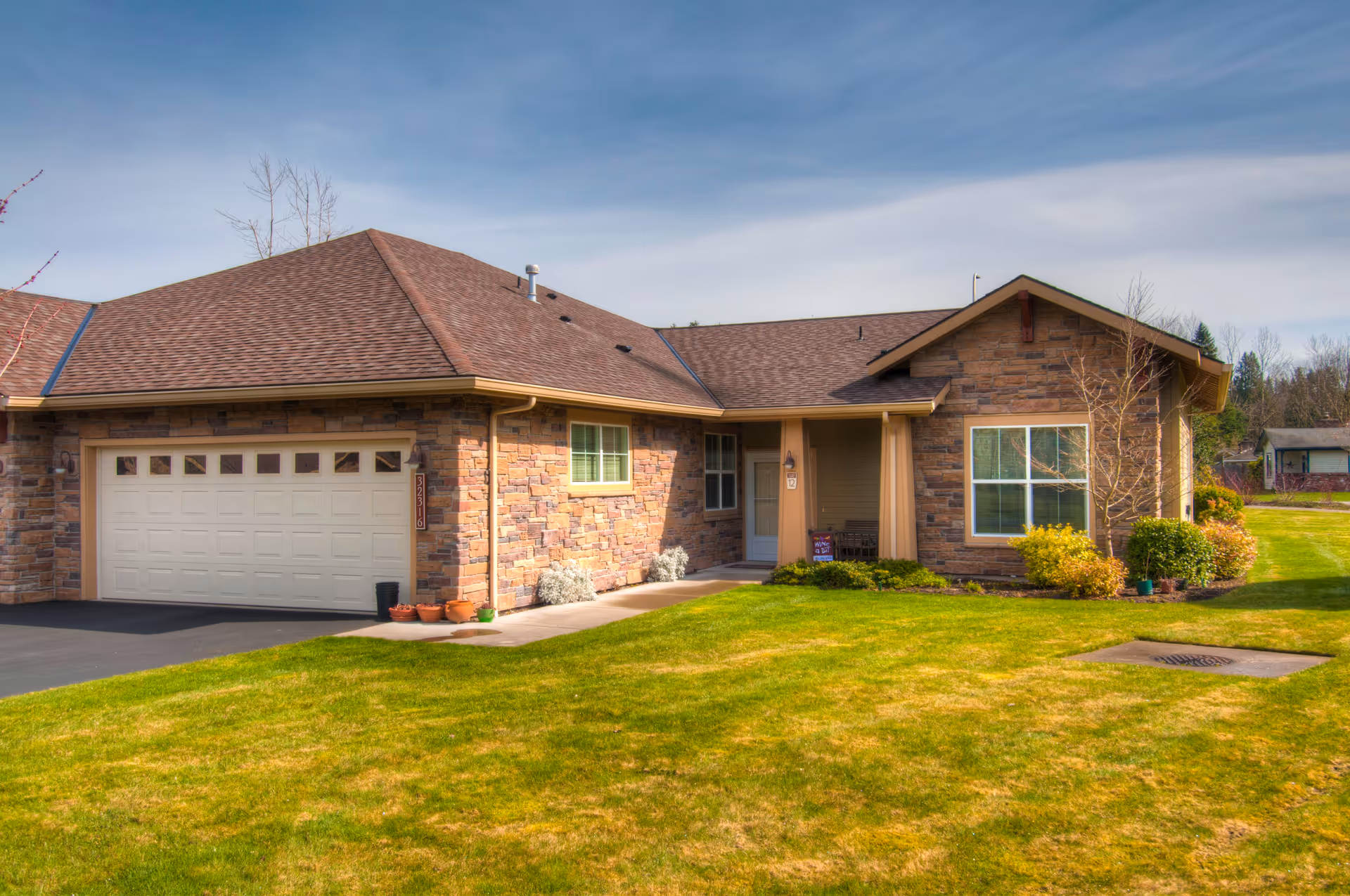 Exterior view of a single-story residential building with a stone facade, a two-car garage, a front porch, and a well-maintained green lawn under a partly cloudy sky.