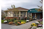 Front exterior of a low-rise senior living building with a covered entrance awning and landscaped flower beds.