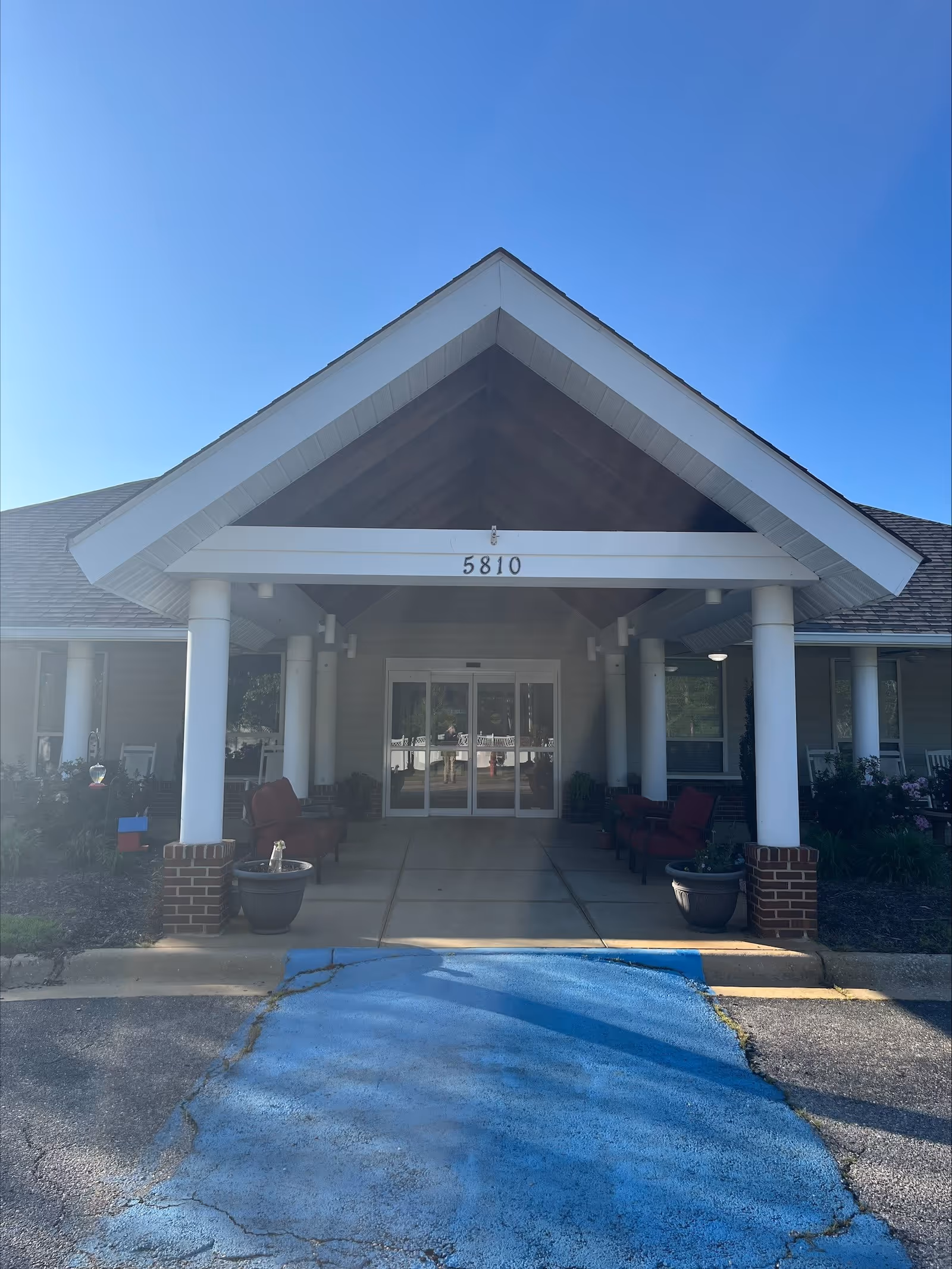 Entrance of a building with a covered porch supported by white columns and a peaked roof. The building has the number 5810 displayed above the entrance doors. There are red cushioned chairs and potted plants on either side of the entrance. The sky is clear and blue.