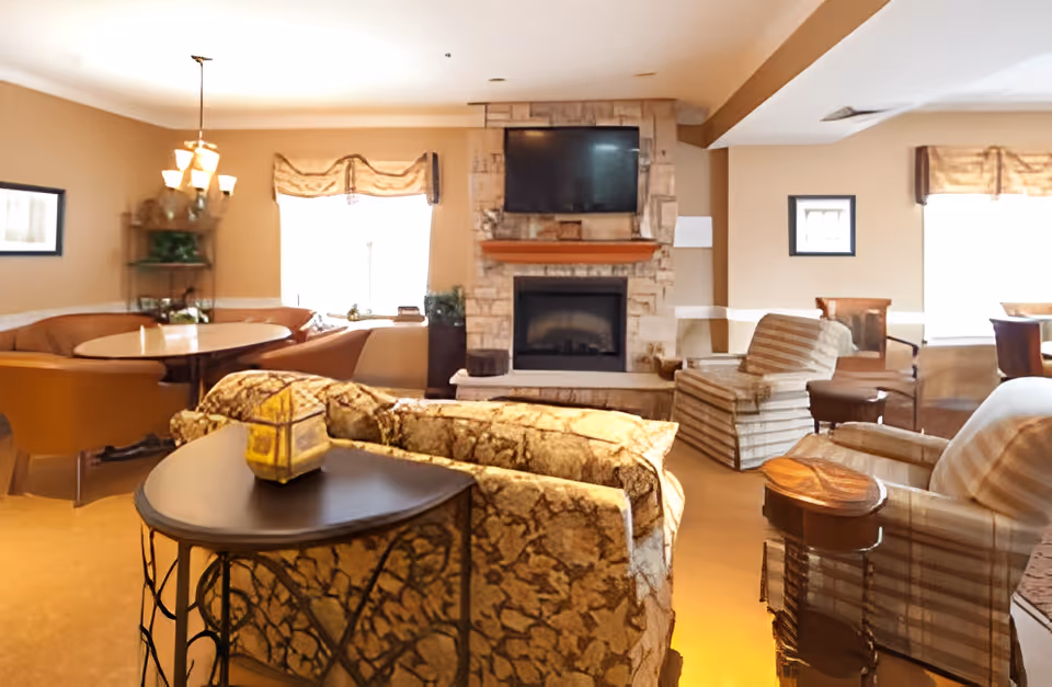 Cozy communal living room with sofas and armchairs facing a stone fireplace with a mounted TV, plus dining tables by the windows.