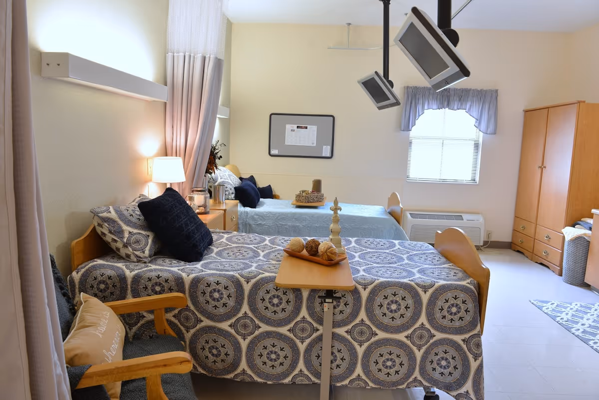 A bright and clean bedroom in a senior living facility with two single beds covered in patterned bedspreads. Each bed has pillows and a small bedside table with a lamp. There is a wooden chair with a cushion, a small overbed table with decorative items, two ceiling-mounted televisions, a window with a curtain, and a wooden wardrobe. The room has light-colored walls and flooring.