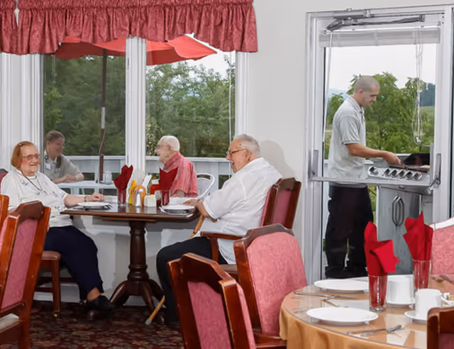Several elderly residents sit around a dining table in a bright dining room while a staff member grills outside on a patio.
