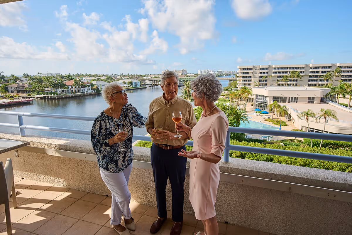 Three elderly people standing on a balcony overlooking a waterfront with buildings and palm trees in the background, holding drinks and engaging in conversation on a sunny day.
