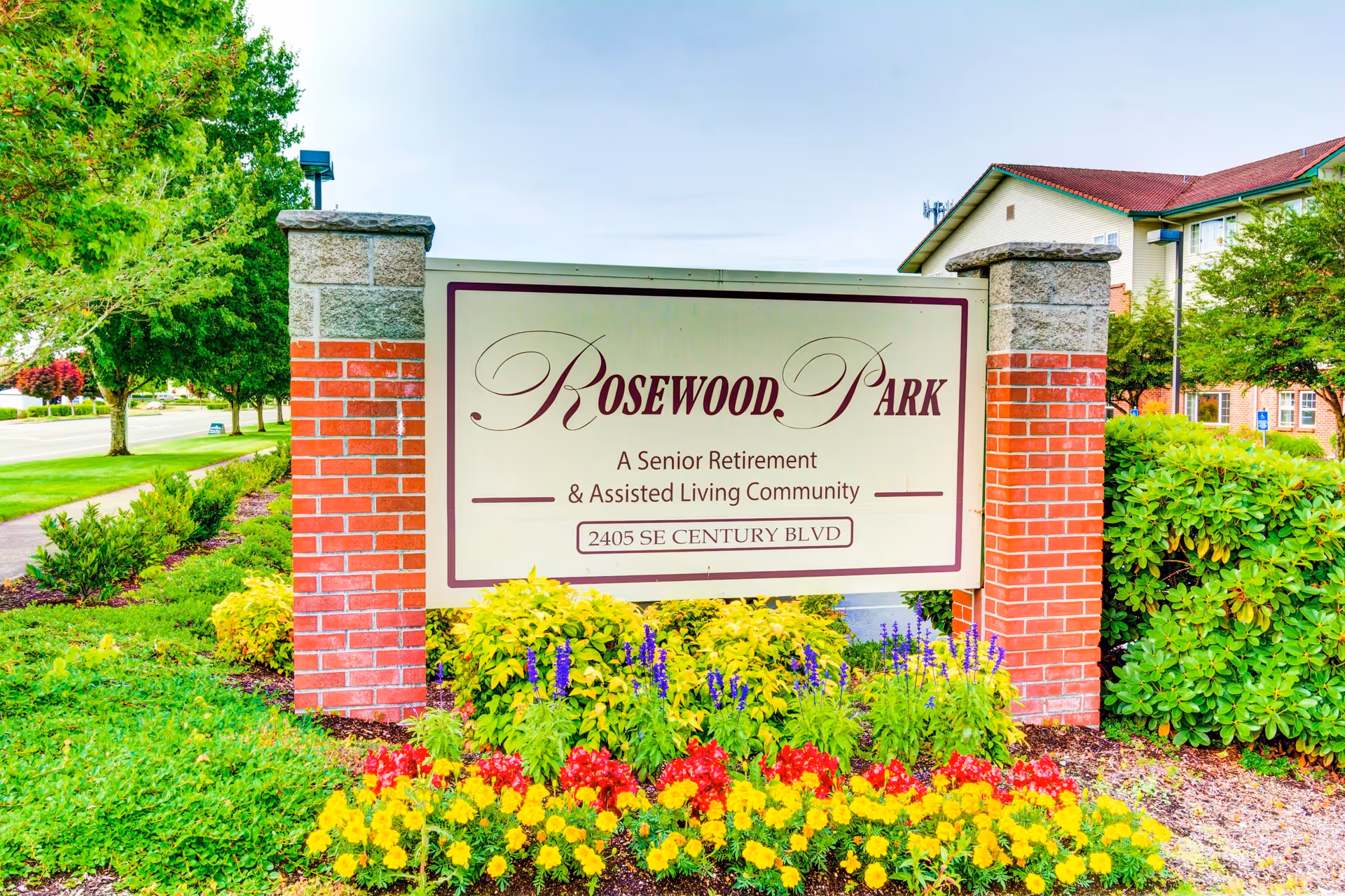 Entrance sign for Rosewood Park, a senior retirement and assisted living community, surrounded by colorful flowers and greenery with a building and trees in the background.