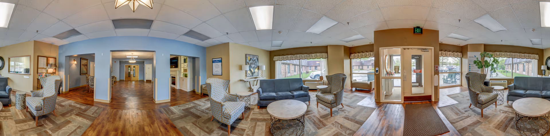 Panoramic view of a senior living facility common area with multiple seating arrangements including armchairs and sofas, large windows letting in natural light, wooden and patterned flooring, and an entrance door with glass panels. The space is decorated with framed pictures, plants, and soft lighting fixtures.