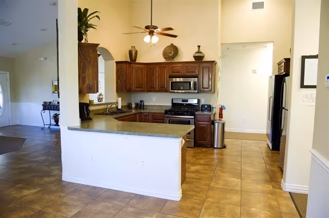 Interior view of a kitchen area in an assisted living facility featuring wooden cabinets, a microwave, a stove, a refrigerator, a ceiling fan with lights, and a countertop with a greenish surface. The floor is tiled, and there is an open space leading to other parts of the facility.