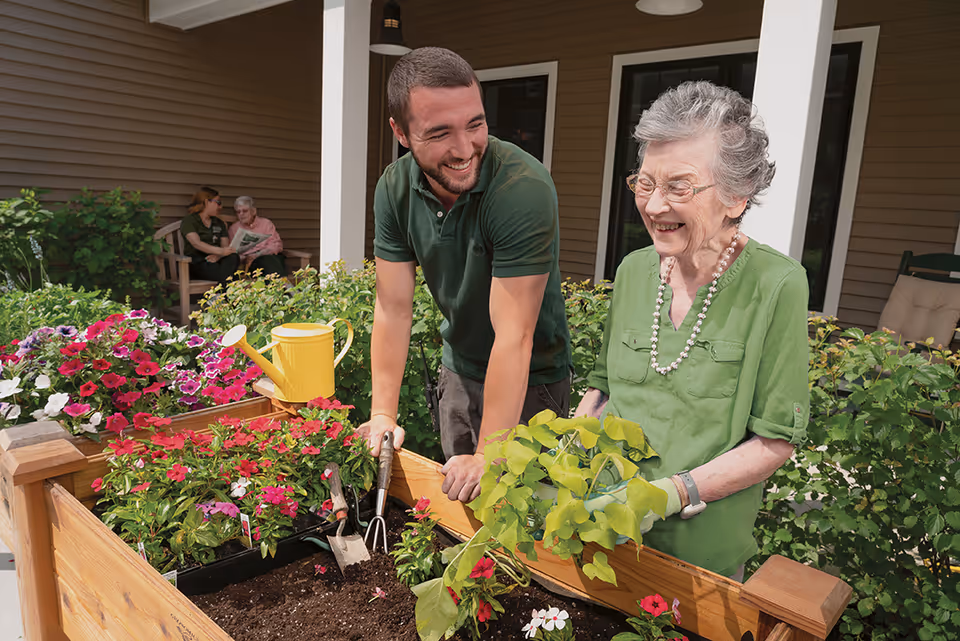 An elderly woman and a younger man are gardening together at a raised wooden garden bed filled with flowers and plants. The woman is holding a green leafy plant and smiling, while the man is leaning on the garden bed and smiling back at her. In the background, two other people are sitting and talking on a bench near the building's entrance.
