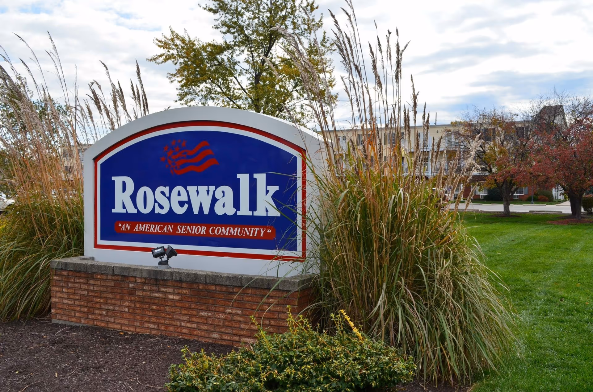 Outdoor view of a large sign for Rosewalk, an American senior community, surrounded by tall grasses and bushes with a building and trees in the background under a cloudy sky.