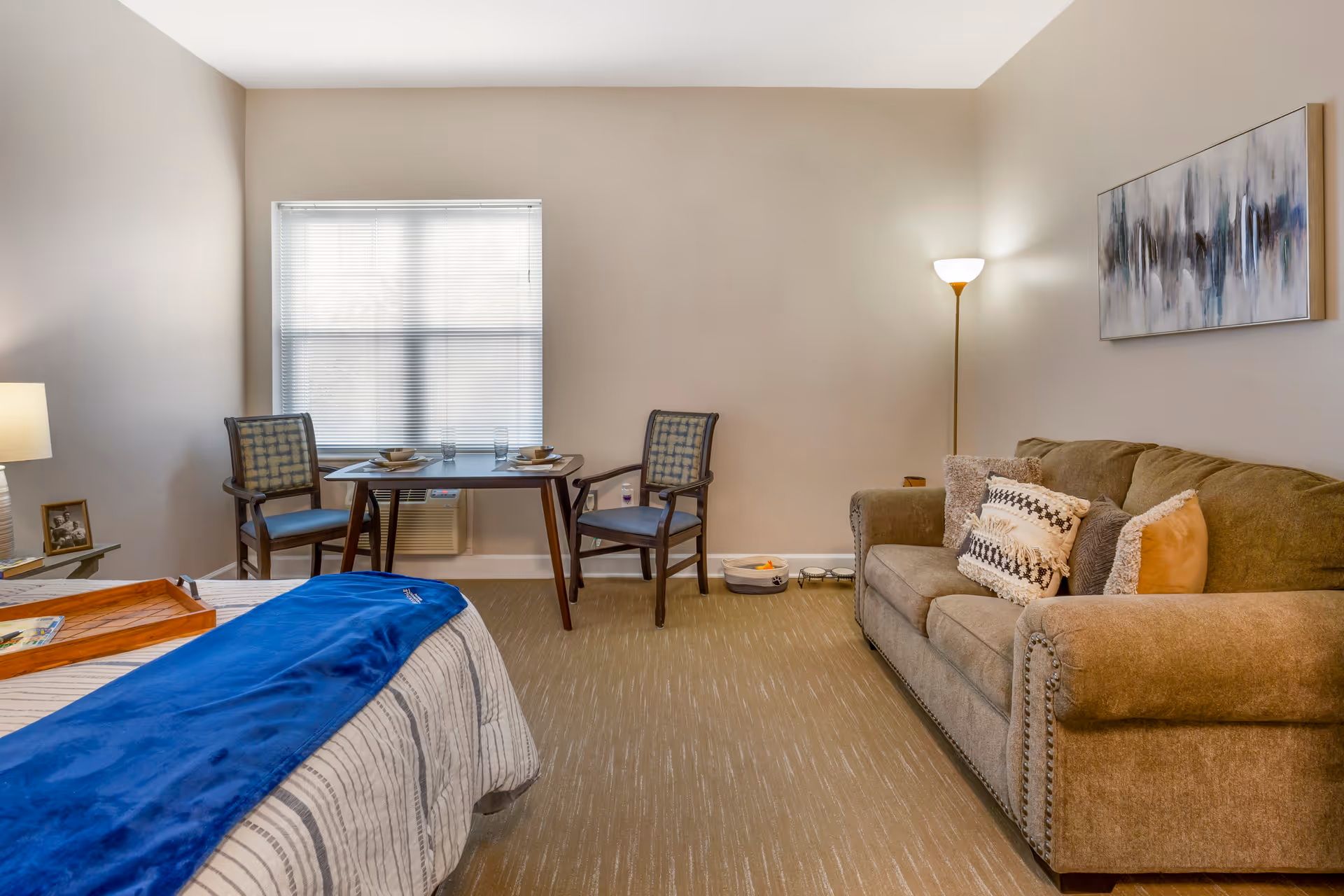 Furnished senior living bedroom with a bed in the foreground, a small table and two chairs by a window, and a sofa with pillows along the right wall.