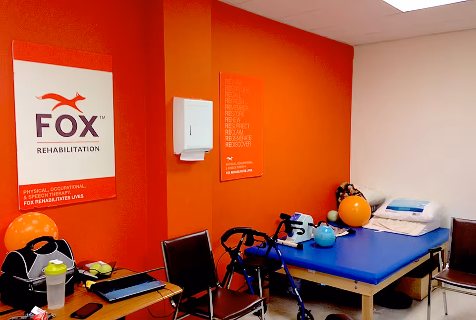 A rehabilitation therapy room with an orange accent wall featuring FOX Rehabilitation posters. The room contains a blue therapy table with pillows and therapy balls, a walker, two chairs, a table with a laptop, water bottle, and other small items.