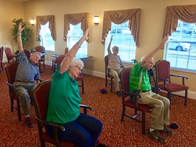 A group of older adults seated on chairs in a carpeted activity room raising one arm during a seated exercise.