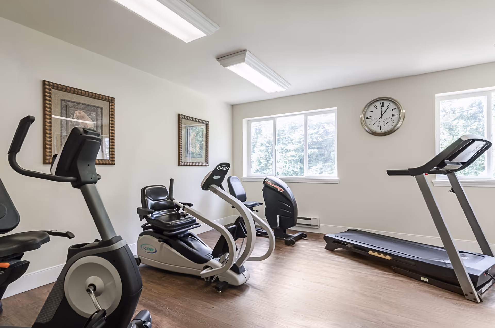 A bright exercise room with large windows, featuring a treadmill, two stationary bikes, and framed artwork on the walls. A clock is mounted on the wall between the windows.