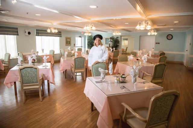 A bright dining room with pink-tablecloth-covered tables and a uniformed staff member standing near the center.