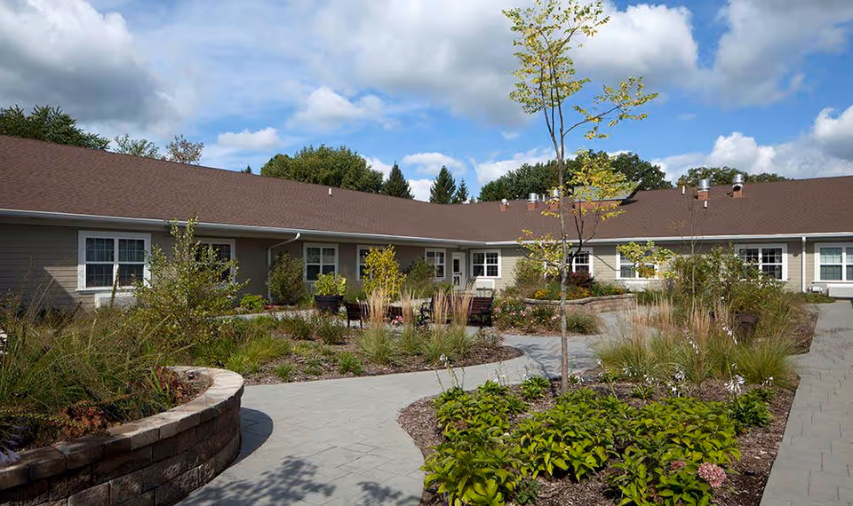 Outdoor courtyard area of The Laurel at Vernon Hills Memory Care facility featuring a paved walkway, landscaped garden beds with various plants and small trees, and a single-story building with multiple windows surrounding the courtyard under a partly cloudy sky.