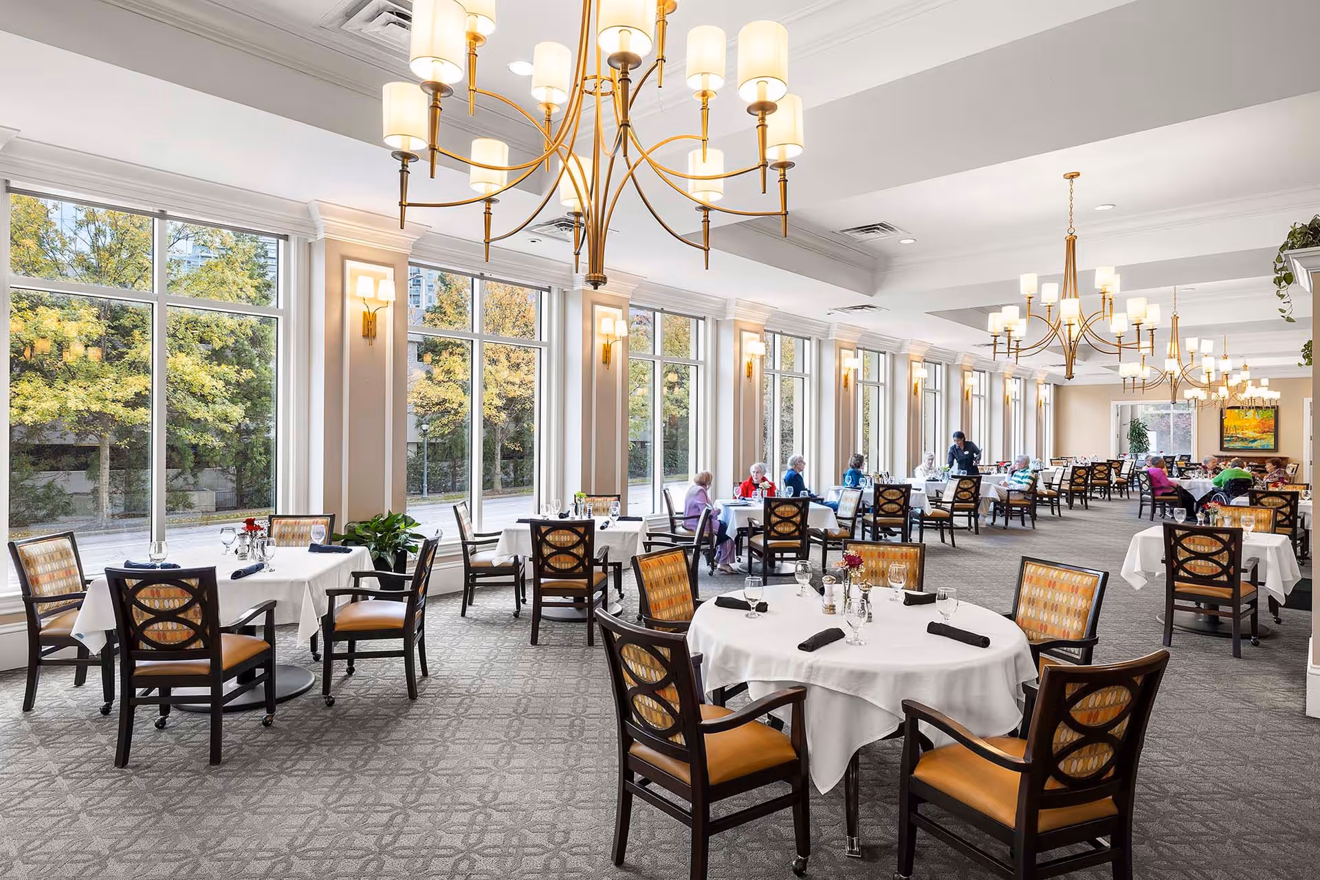 A spacious, well-lit dining room with large windows showing outdoor greenery. The room features round and square tables covered with white tablecloths, set with glassware and napkins. Several elderly people are seated at tables, and a staff member is attending to them. Elegant chandeliers hang from the ceiling, and the room has a warm, inviting atmosphere.