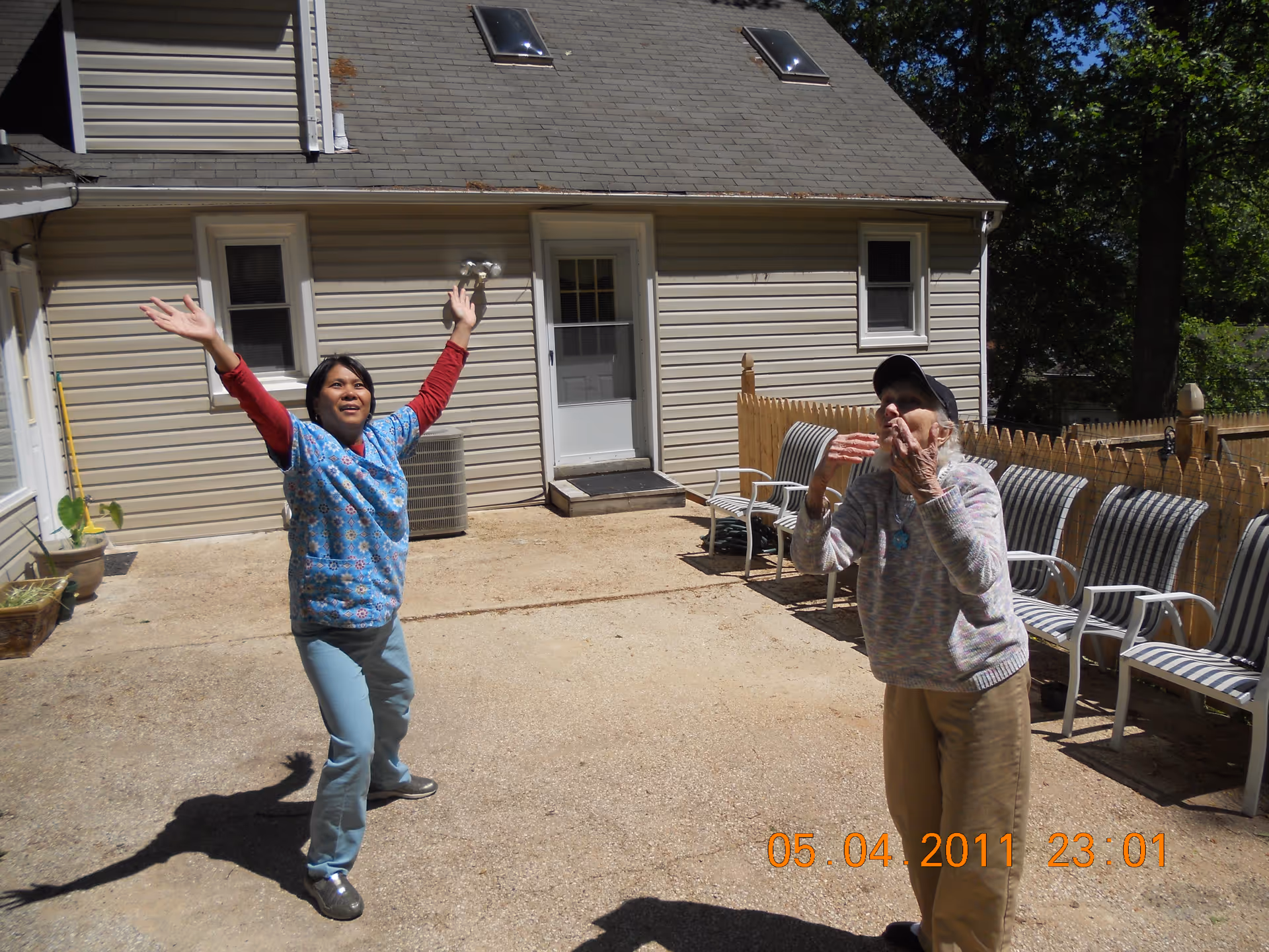 Two women are outside in a fenced patio area in front of a beige house. One woman, wearing a blue patterned shirt and light blue pants, has her arms raised and is looking up. The other woman, wearing a gray sweater, beige pants, and a black cap, is also looking up with her hands raised near her face. Several striped chairs are lined up against the fence on the right side.