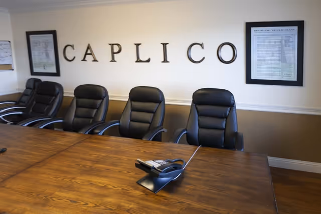 A conference room with a long wooden table and six black leather office chairs. On the table is a conference phone. The wall behind the chairs has large letters spelling 'CAPLICO' and two framed documents on either side.