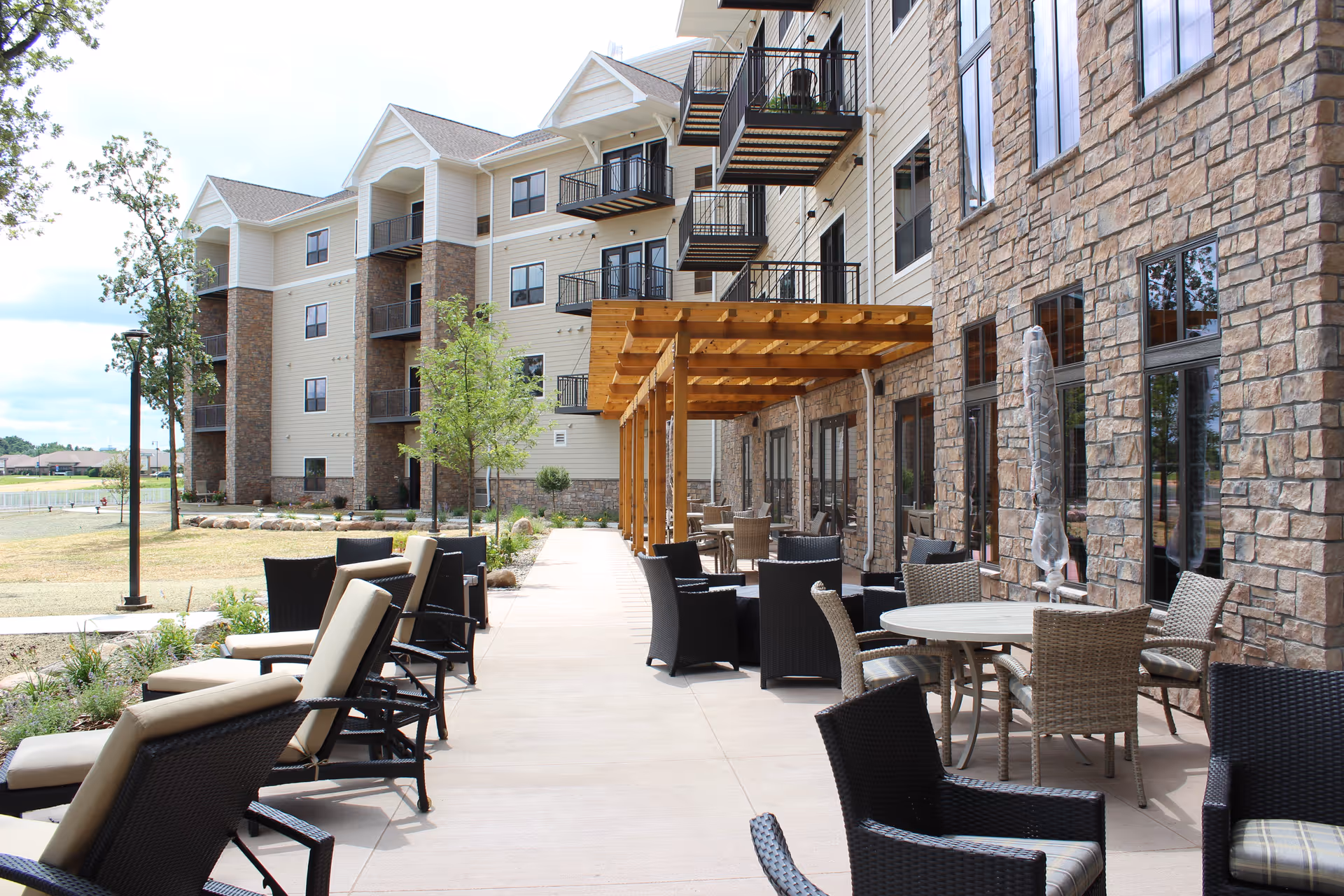 Outdoor patio area with wicker chairs, tables and lounge seating beside a multi-story stone and siding senior living building with balconies.