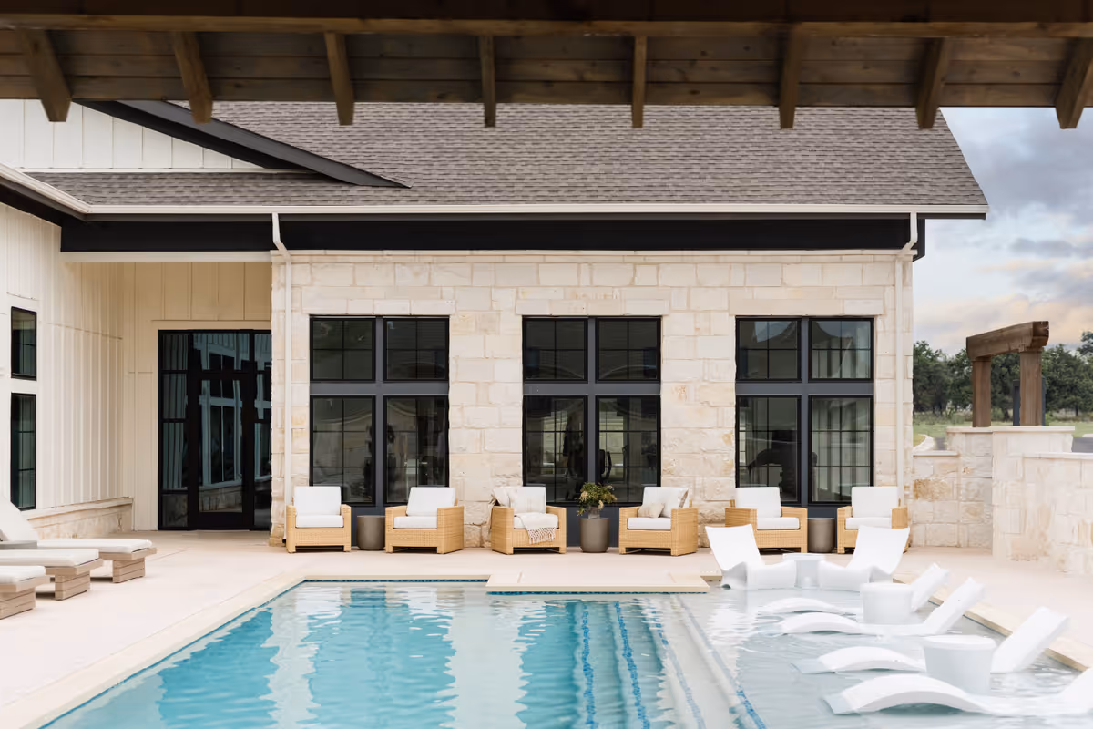 Outdoor pool area at Woodland Cottages Senior Living Fredericksburg featuring a rectangular swimming pool with built-in lounge chairs partially submerged in water. Surrounding the pool are cushioned wicker armchairs and lounge chairs, set against a stone building with large black-framed windows and a covered patio roof.