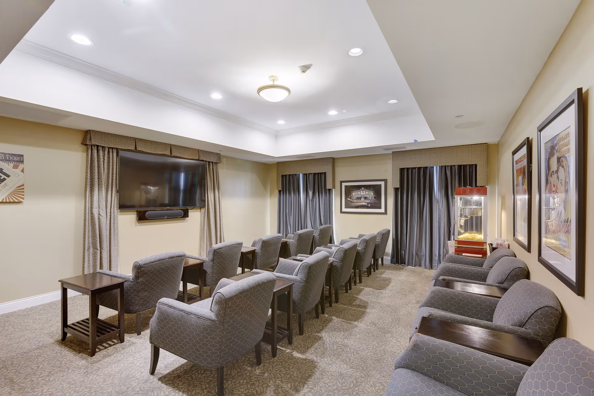 A cozy media room with two rows of gray upholstered armchairs facing a large flat-screen TV mounted on the wall. The room has beige walls, carpeted floor, and ceiling lights. There are framed pictures on the walls and a popcorn machine in the corner near the window with dark curtains.