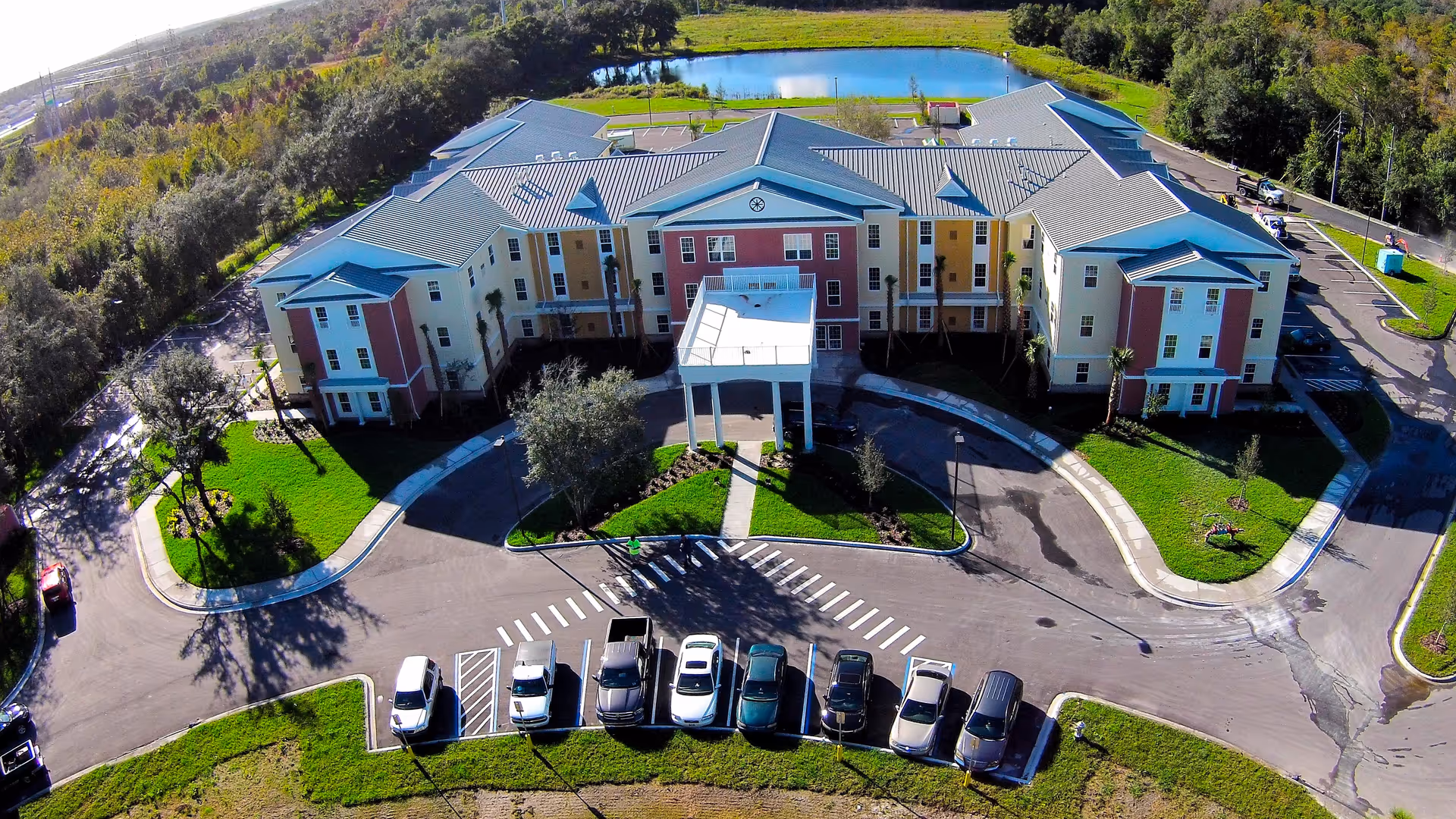 Aerial view of a large, three-story senior living facility building with a covered entrance and a semicircular driveway. Several cars are parked in front, and the building is surrounded by green lawns, trees, and a pond in the background.