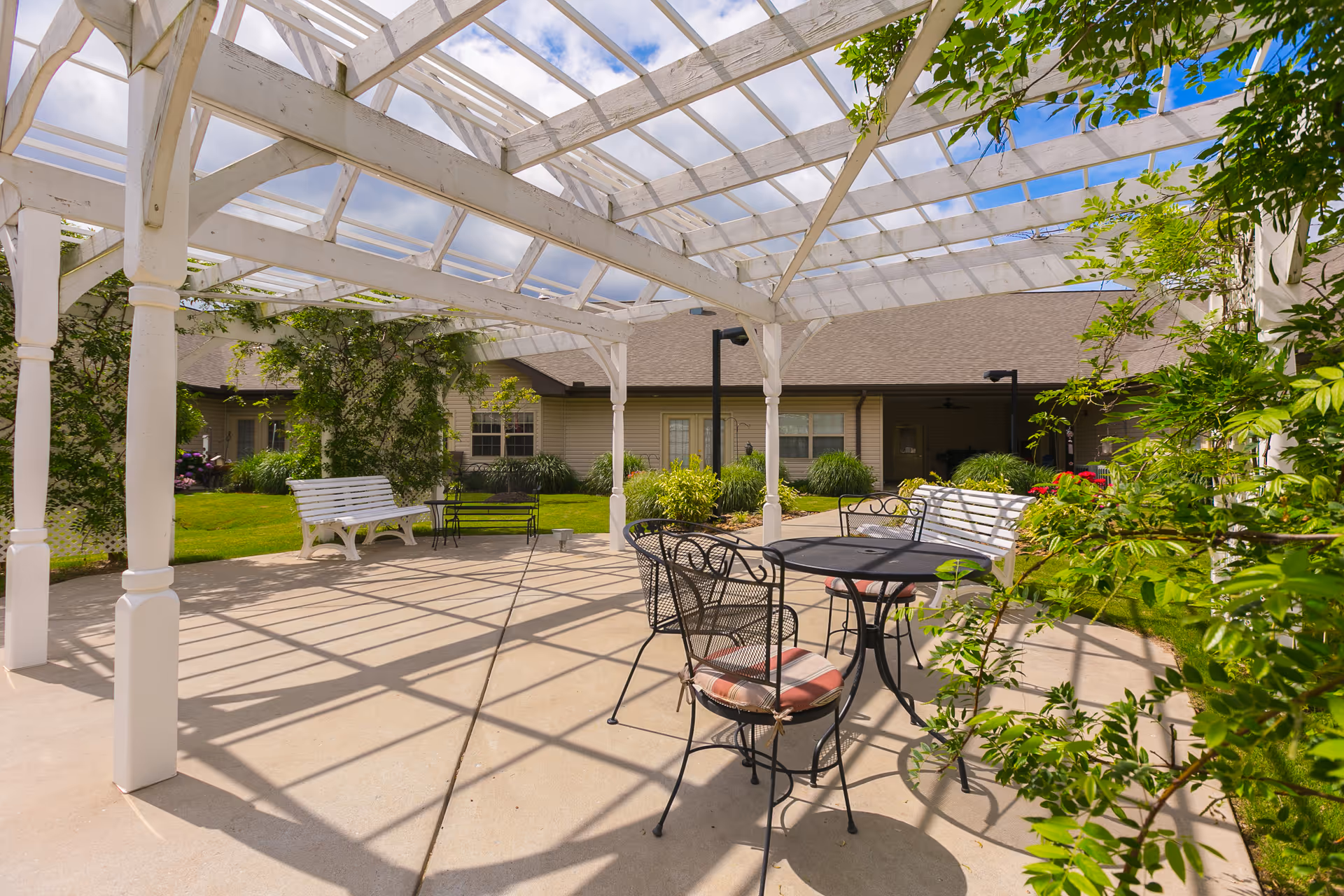 Outdoor patio area with a white pergola casting shadows on the concrete floor. There are white benches and black metal chairs with a table, surrounded by green plants and shrubs. A building with beige siding and windows is visible in the background under a partly cloudy sky.