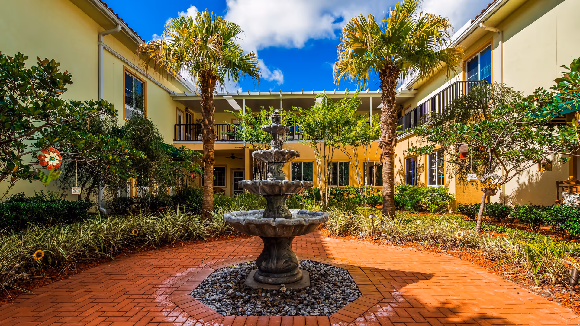 A sunny courtyard at The Windsor at San Pablo featuring a three-tiered stone fountain in the center, surrounded by palm trees, shrubs, and a brick-paved walkway. The courtyard is enclosed by a two-story building with balconies and windows, under a bright blue sky with some clouds.
