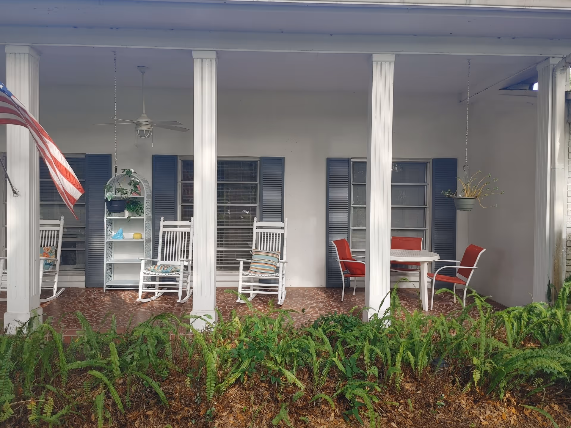 Front porch of a building with white pillars, two white rocking chairs with striped cushions, a white shelf with plants and decorations, a round white table with four red cushioned chairs, hanging plants, and an American flag on the left side. There are blue shutters on the windows behind the porch and green ferns in front of it.